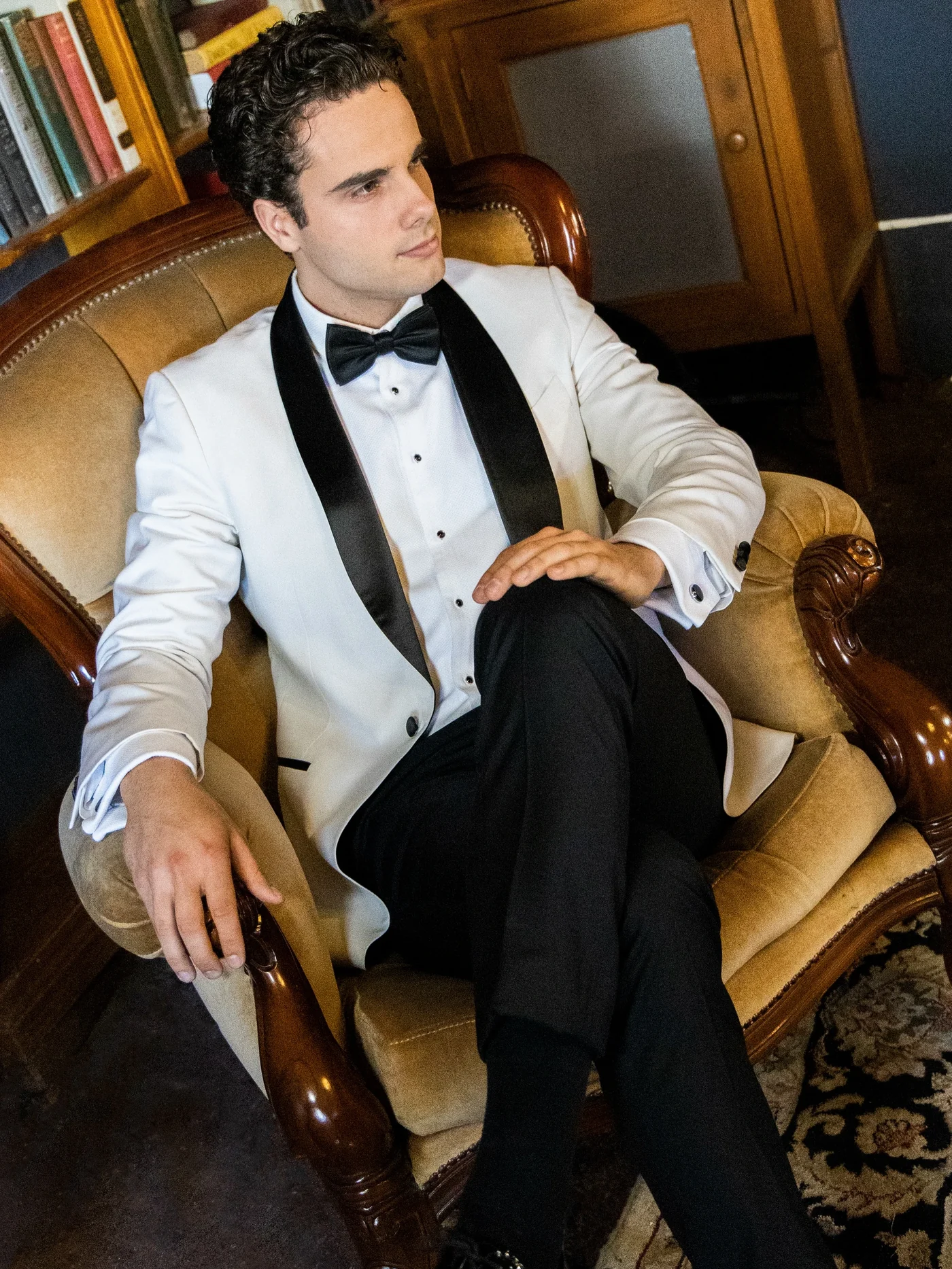 The groom in a white dinner jacket, seated beside the library shelves.